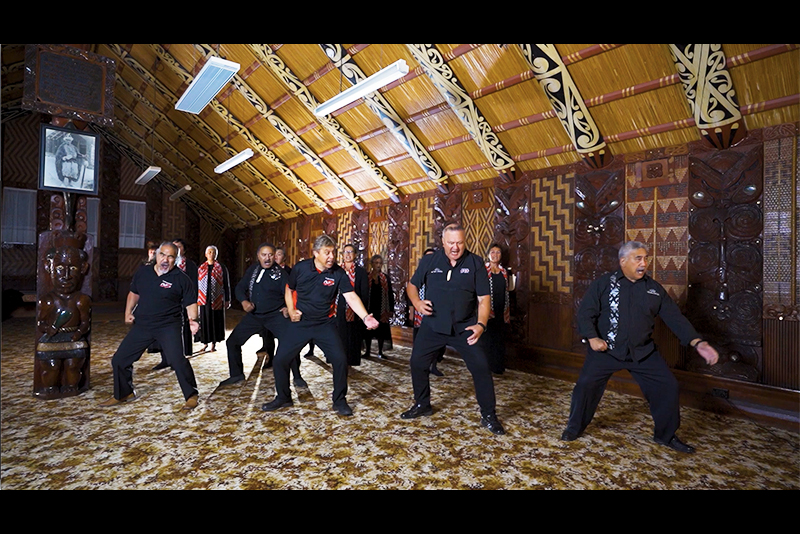 Six men are performing a cermonial dance in a carved meeting hall. There are women singing at the back