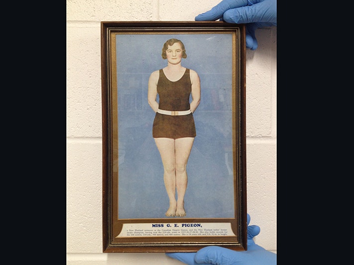 Gladys Pidgeon in her swimming costume. Gifted to Te Papa by Pam Marley, 2018. Te Papa A pair of blue-gloved hands holds a framed colourised photo of a woman in a bathing costume.