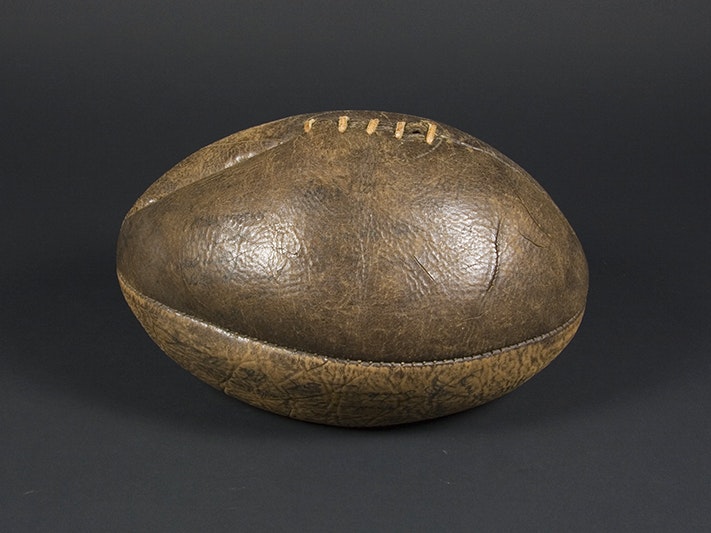 Rugby ball; about 1921; Unknown; leather ball with ink signatures. Purchased 2009. Te Papa (GH012621) A brown leather rugby ball on a black background. The ball is slightly deflated.