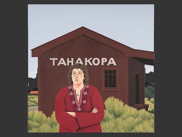 Robin White, Glenda at Tahakopa, oil on canvas, 1978. Painting of a woman standing in front of a rural train station. The station is made of corrugated iron and painted brown. The woman stands with her arms crossed, wearing a red dress with a floral design around its swooping neck line