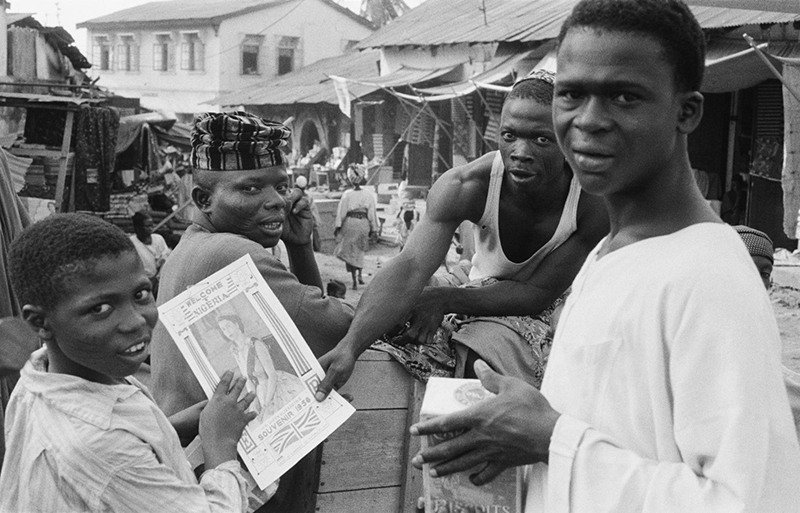 Some young Nigerian men have a magazine with a picture of the Queen on the cover. They are showing it to the photographer