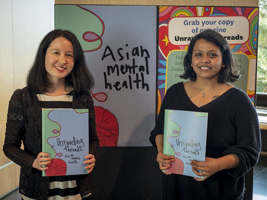 Grace Gassin and Mehwish Mughal, 2022. Photo by Daniel Crichton-Rouse. Te Papa Grace and Mehwish stand in front of a sign advertising the Asian Mental Health project, holding copies of the zine