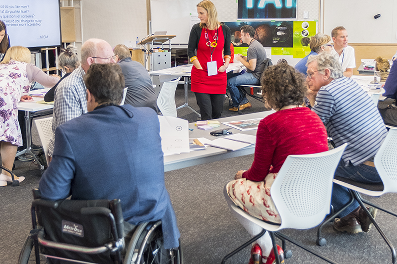 A room where people are sitting at tables in a workshop. One person is sitting in a wheelchair