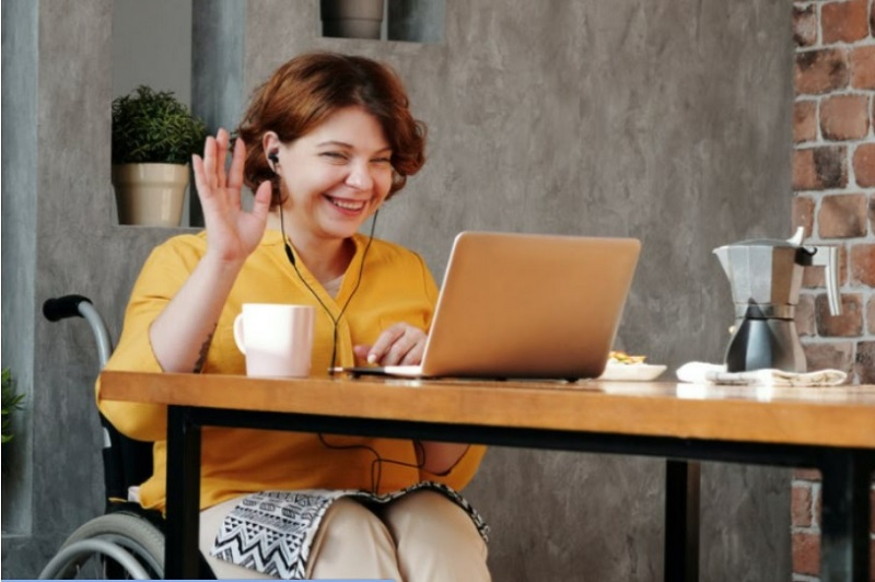 A woman sits in her wheelchair at a table, facing a laptop and waving at the screen with a smile on her face