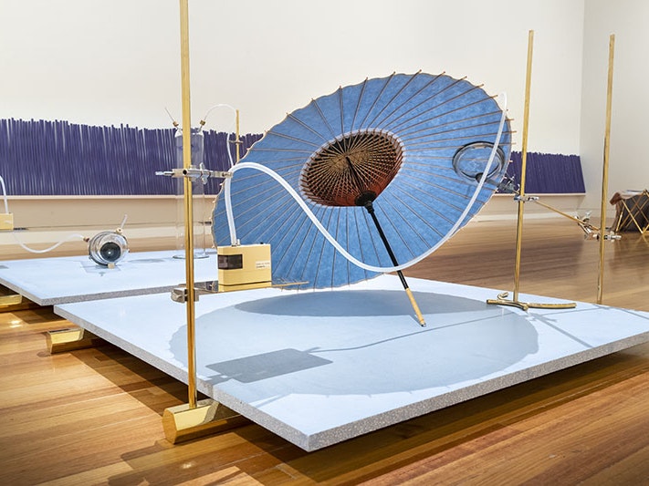 Dane Mitchell, Iris, Iris, Iris (detail), 2017. Te Papa (2021-0027-1) A platform sits on gold-coloured hexagonal feet. On the platform rests an iris-coloured Japanese umbrella, a camera lens and an iris flower in seperate glass jars. In the background a row of large purple incense sticks against the wall.