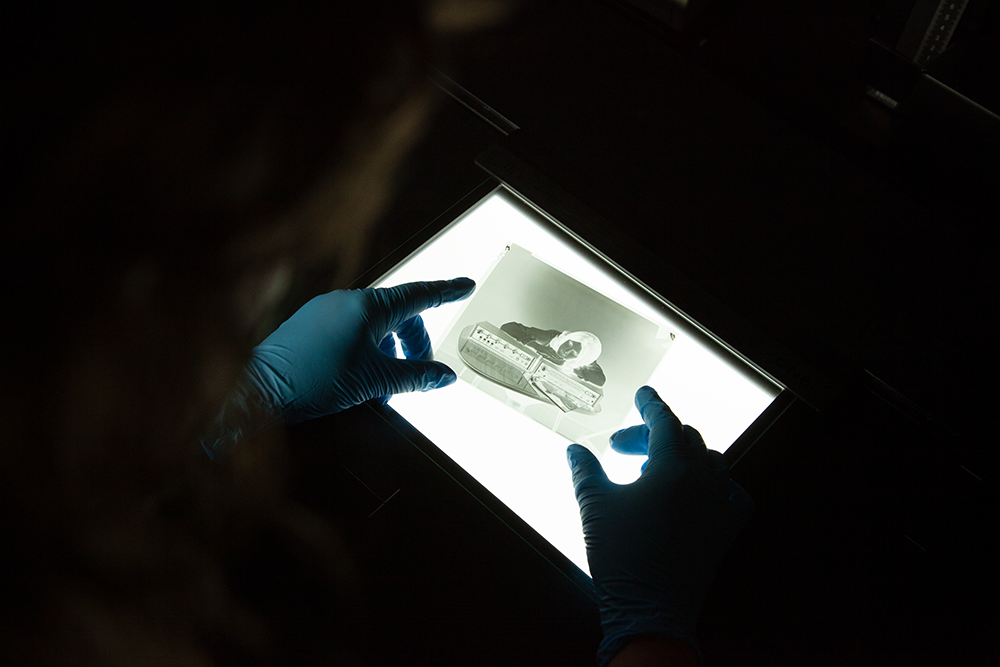 A dark photo of two hands wearing blue gloves holding a photographic negative on a lightbox. The negative shows a woman leaning on a piece of machinery