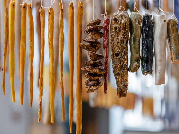 Installation view of Kate Newby, SHE’S TALKING TO THE WALL, 2012-2021 in Kate Newby: YES TOMORROW at Te Pātaka Toi Adam Art Gallery, 2021. Photo by Ted Whittaker and courtesy of Te Pātaka Toi Adam Art Gallery Multiple ceramic pieces hang by string, some are long and thin and dyed an orange colour; others are a variety of shapes and colours – white, dark blue, green, brown, shorter, squatter, and some have holes in them
