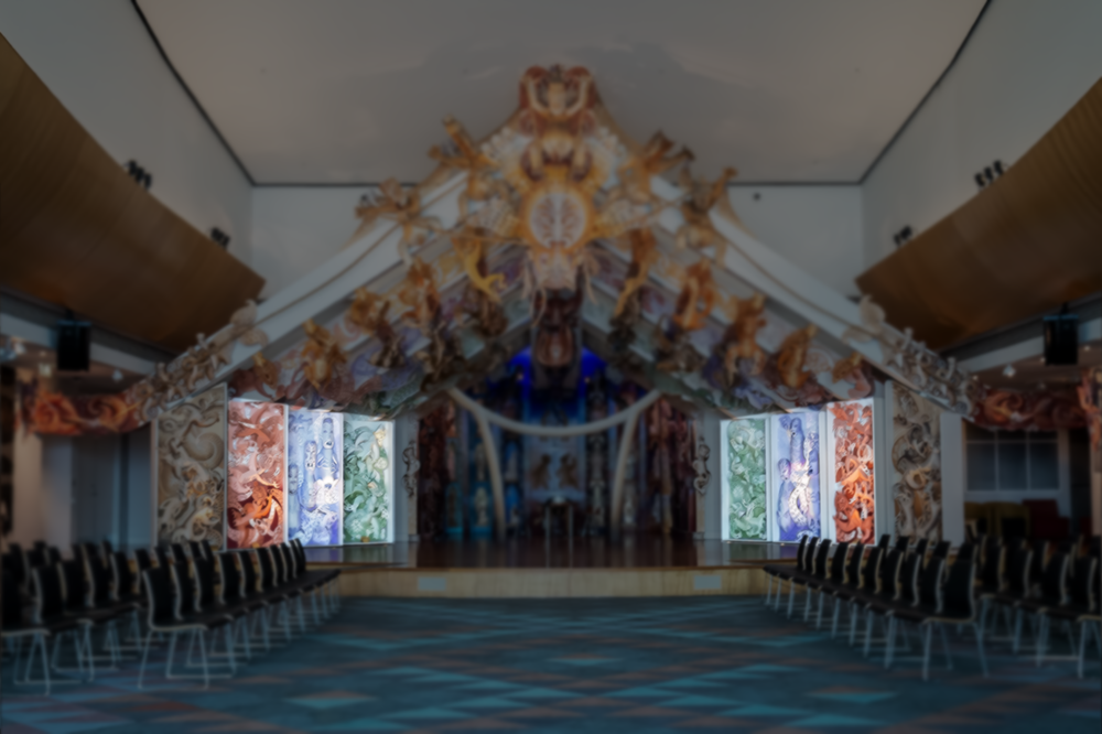 A multicoloured stage with an open-wall house at the back with carvings made of MDF on the sides and rafters. There are chairs either side of the stage set out so they face each other.  One section is highlighted the rest of the image is darkened.