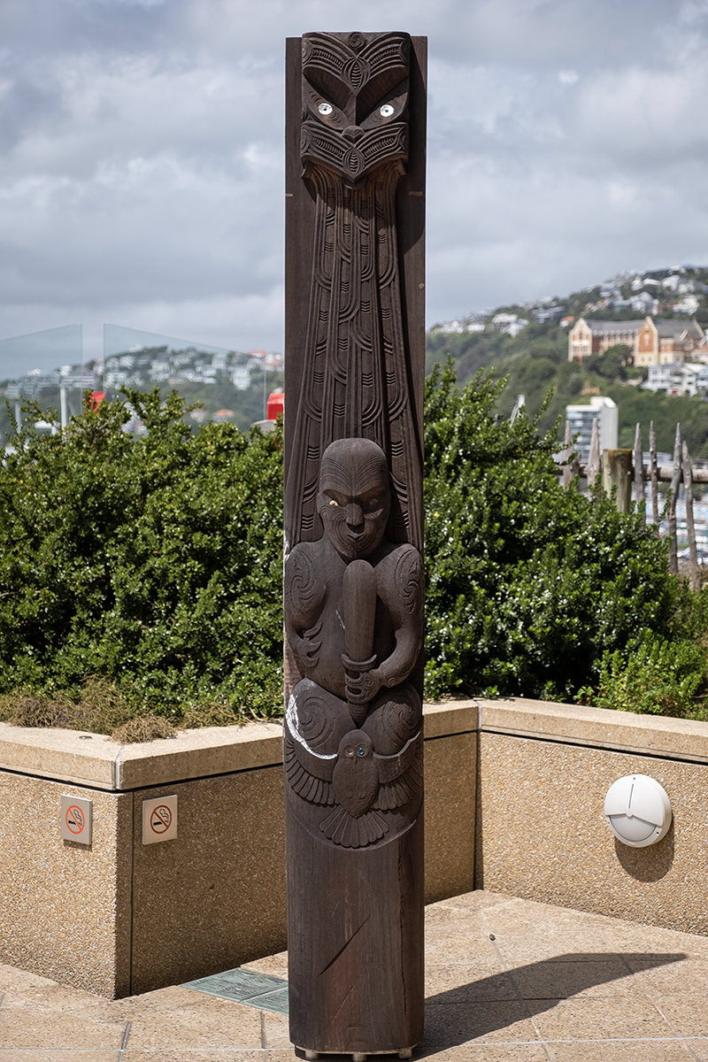 A carved piece of wood standing upright on a balcony. There's a blue sky behind it and a glass fence.