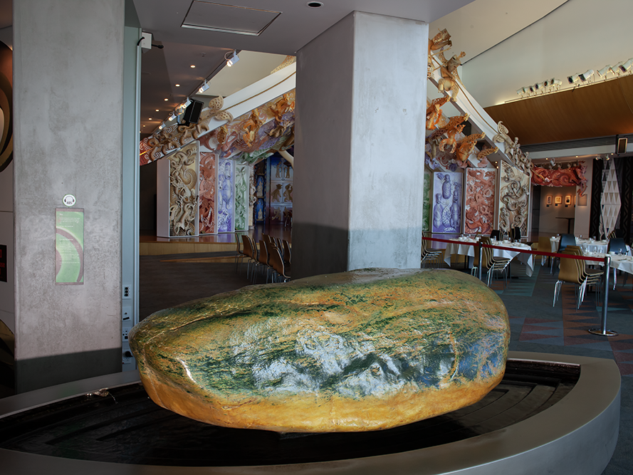 pounamu_kaitiaki_tehono-20.png A large boulder in a room. It is partly green and partly yellow. It's sitting in a large wet bowl with running water.