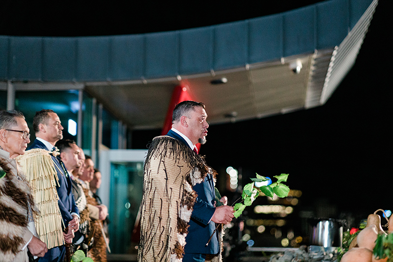 A man holding a green leafy branch is chanting outside in the early morning. There are several men standing behind him.