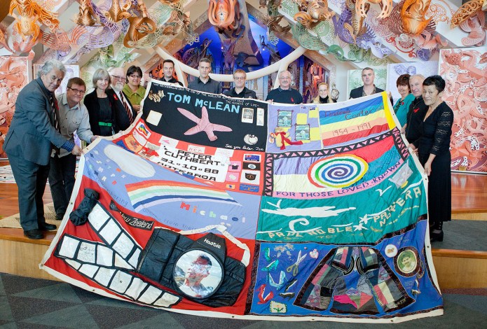 Thirteen people hold up a block from the New Zealand AIDS Memorial Quilt to face the camera, in Te Papa’s marae