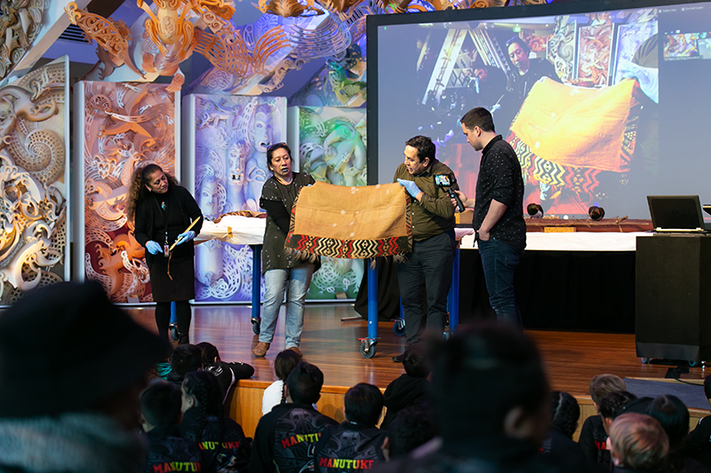 Four people are standing on a stage. Two are holding a cloak made of feathers, one is talking about the cloak and one is holding a camera. There are school children in the audience.