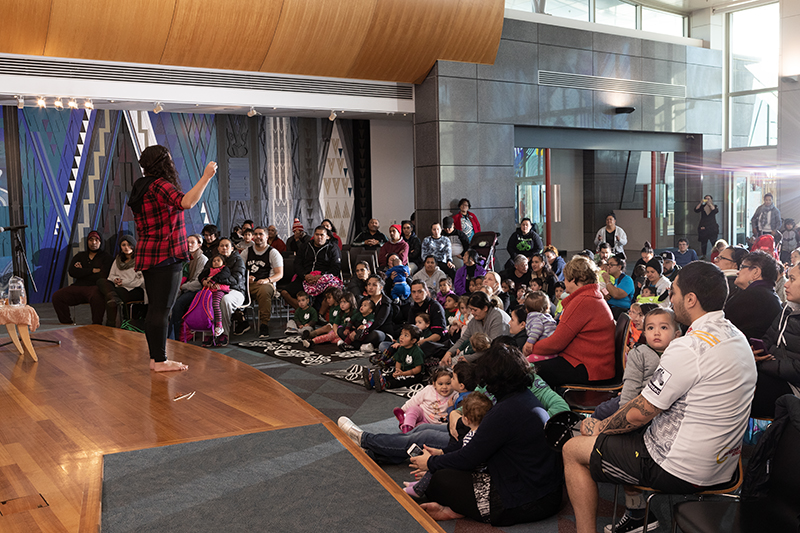 A woman standing on a stage with a lot of parents and children sitting on the floor in front of her.