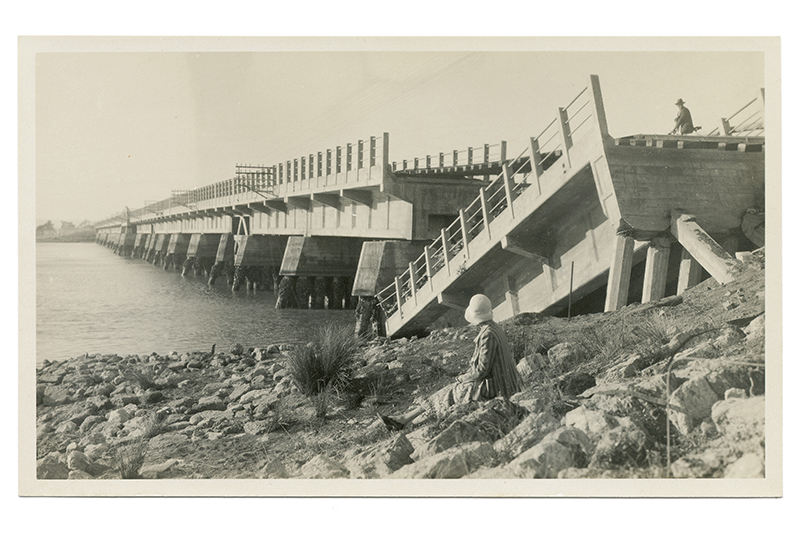 A woman sits on the side of the river looking at a bridge that has collapsed after an earthquake.