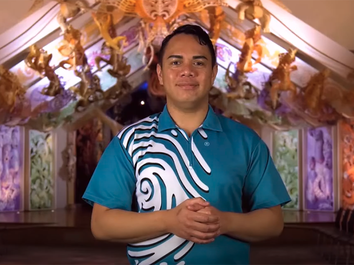 NZSL Introduction to Rongomaraeroa, 2021. Te Papa (168115)   Sponsored by the New Zealand Sign Language Fund A man standing in front of a carved building with his hands clasped in front of him.