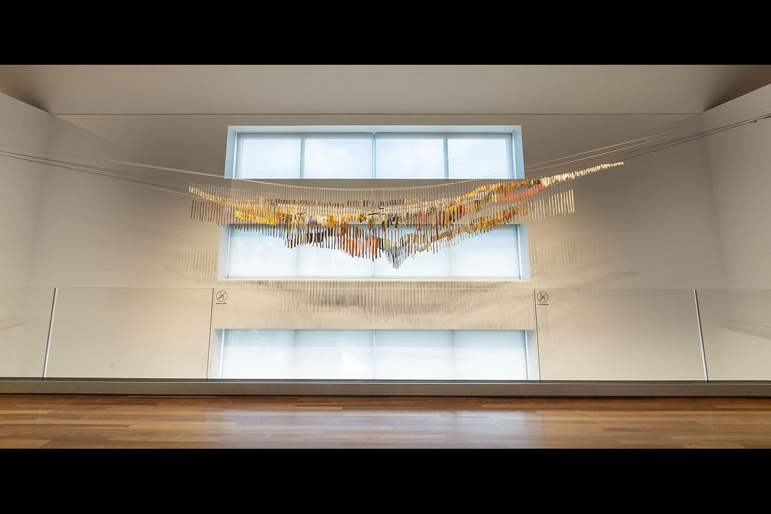 Low-angle photo looking up at three horizontal strips of windows on the wall. In front hangs an artwork consisting of hundreds of sculptures hanging on lines