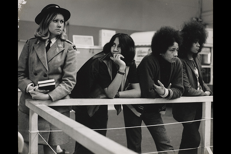 View of three young Māori men leaning on a wooden rail. On their left stands a young blonde woman in a St Johns uniform.