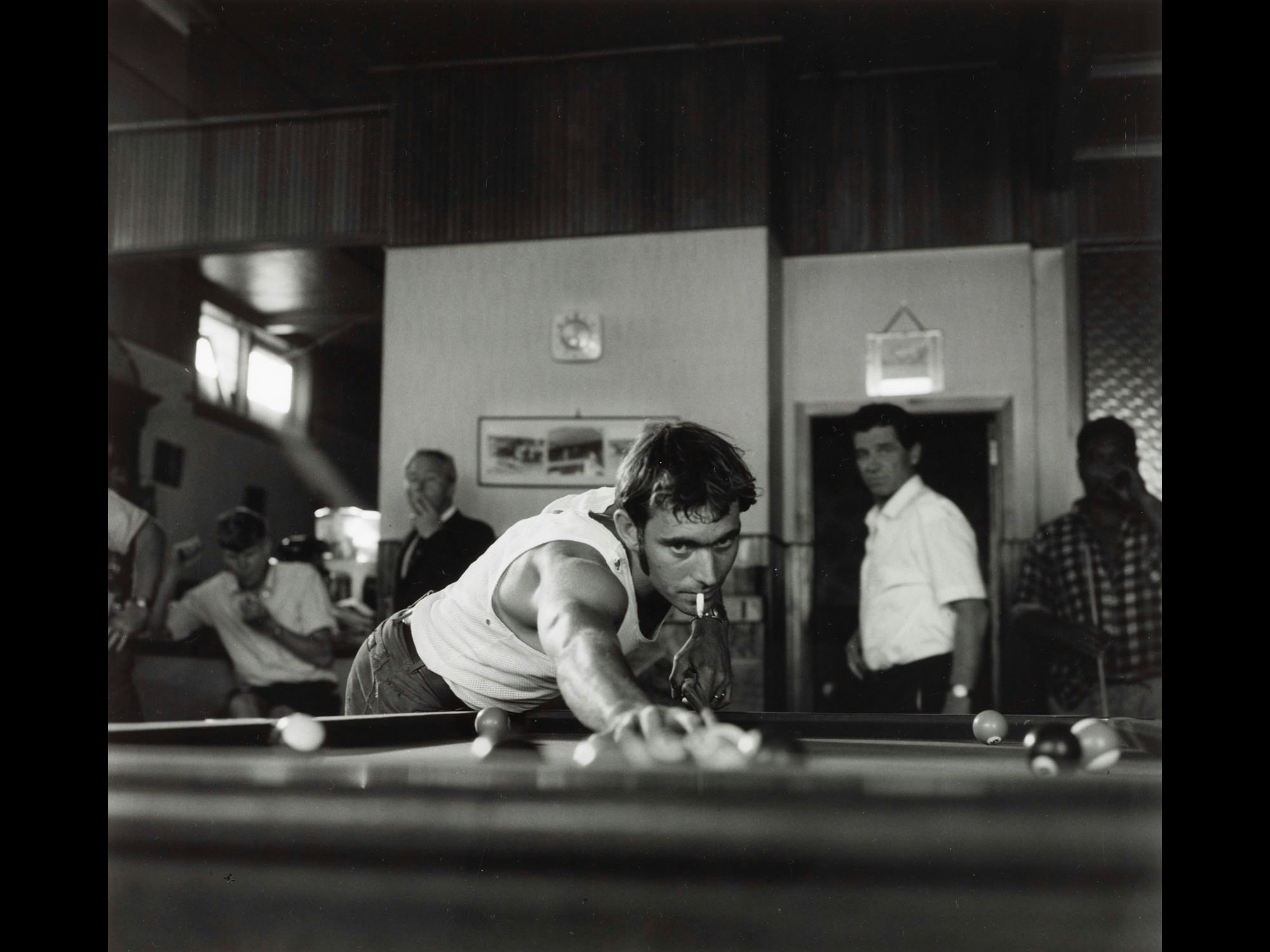 Low-angle shot of a man with a cigarette in his mouth playing pool while other men stand around behind him