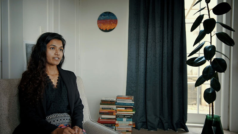 A woman sits on a light grey chair, talking to somewhere out of the frame on the right. Behind her sits two piles of books, and a painting on the wall