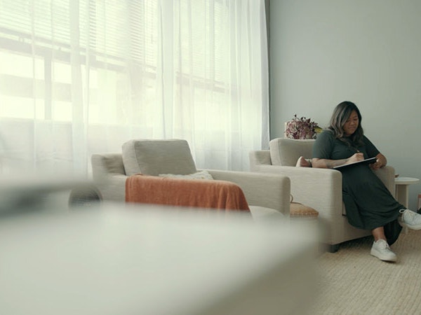 Maria Milmine, 2022. Photo by Julie Zhu. Te Papa A woman sits on a lounge chair, writing on a pad. She is in a well-lit spacious room that has a relaxing feel to it
