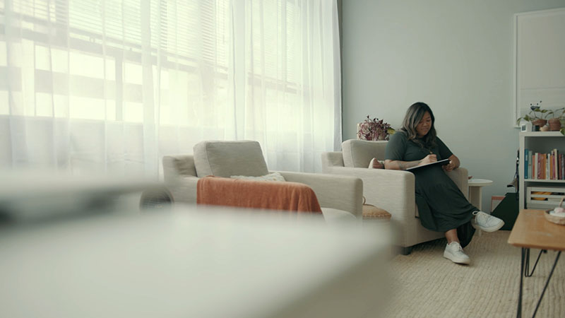 A woman sits on a lounge chair, writing on a pad. She is in a well-lit spacious room that has a relaxing feel to it