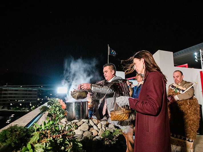 Rangi Matamua and Jacinda Ardern at the Matariki hautapu ceremony, 2022. Te Papa A man and a woman stand outside. It’s early morning and the sky is still dark. The man lifts food out of the pot, releasing steam.
