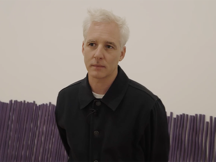 Still from Dane Mitchell, ‘Iris, Iris, Iris’. Te Papa A man with white hair is standing in a room that has large purple incense sticks lying against the wall.