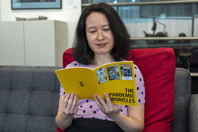 Grace sits on a couch with a large red cushion reading a copy of The Pandemic Chronicles