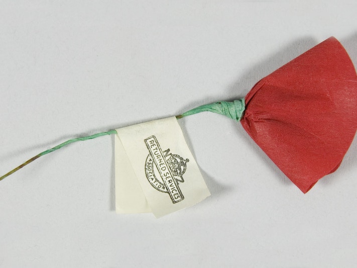 Anzac poppy, New Zealand Returned Services Association; manufacturer(s); 1960s-1970s; paper and wire. Found in collection, 2016. Te Papa (GH025107) A red paper flower representing a poppy with a green stalk made of wire. There is a tag on the stalk with the words Returned Services Association and their emblem.