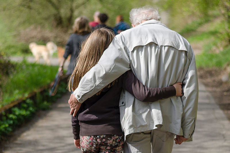 A young girl and an older man – possibly her grandfather – walk arm in arm along a path surrounded by trees