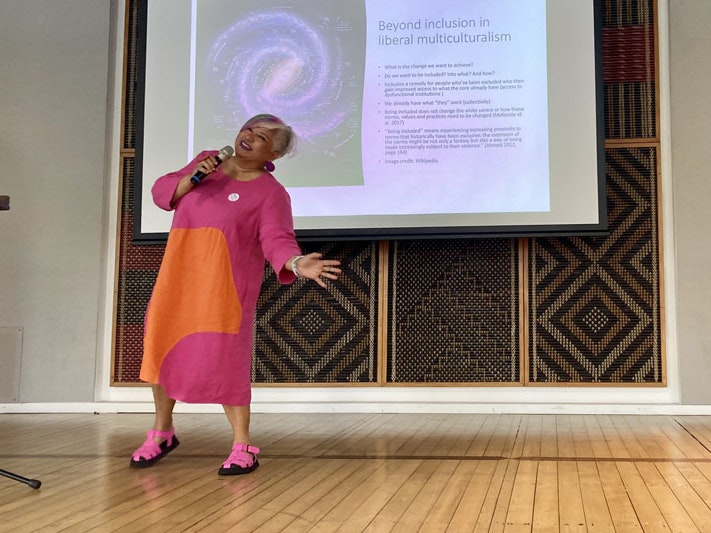 Dr Ruth DeSouza at the Reimagining Asian Mental Health hui, 18 March 2023. Photo by Nishhza Thiruselvam / Twitter A woman in a bright pink and orange dress stands on stage, smiling, addressing a crowd