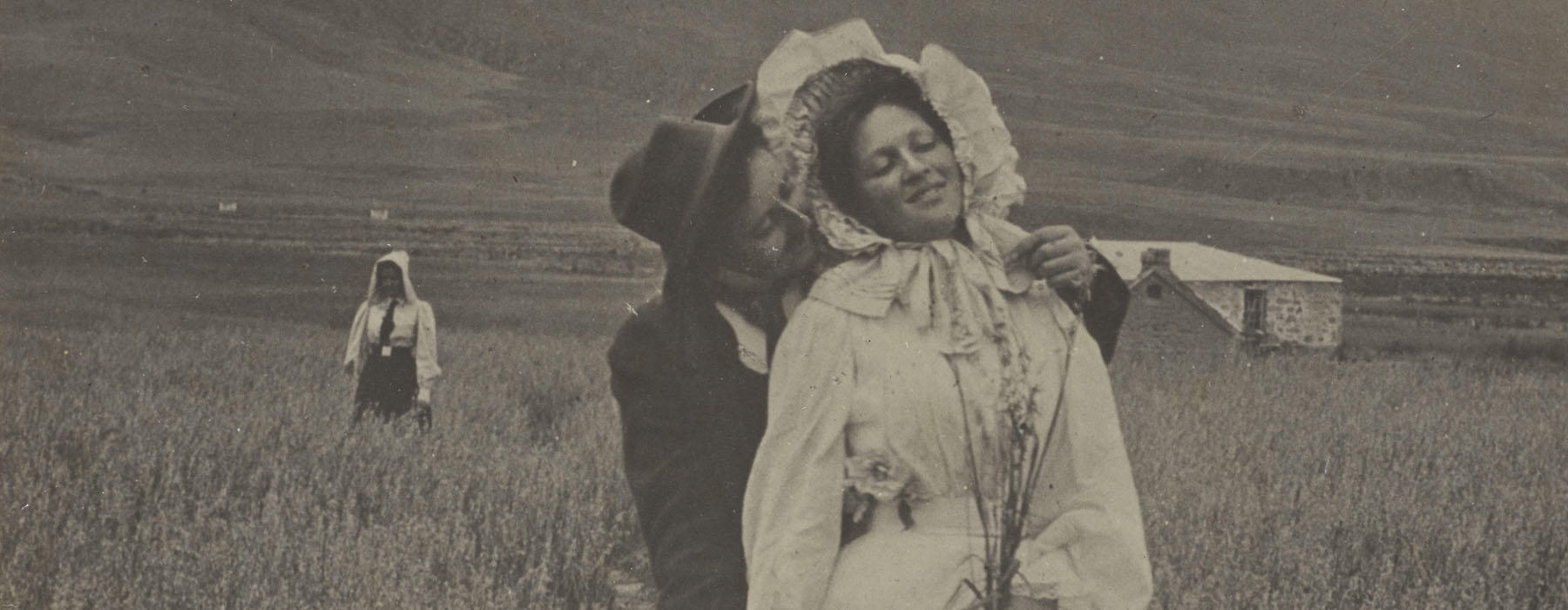 A man hugs a woman from behind in a field. She holds a bunch of flowers. Behind them a woman stands in isolation. Further back, there’s a stone house and hills