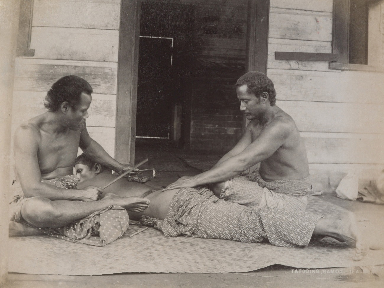 Tattooing, Samoa, 1890-1910, Samoa, by Thomas Andrew. Gift of Alison Beckett and Robert McPherson, 1996. Te Papa (O.001279/01) A sepia photo of two Sāmoan men tattooing another man who is lying down on a flax mat