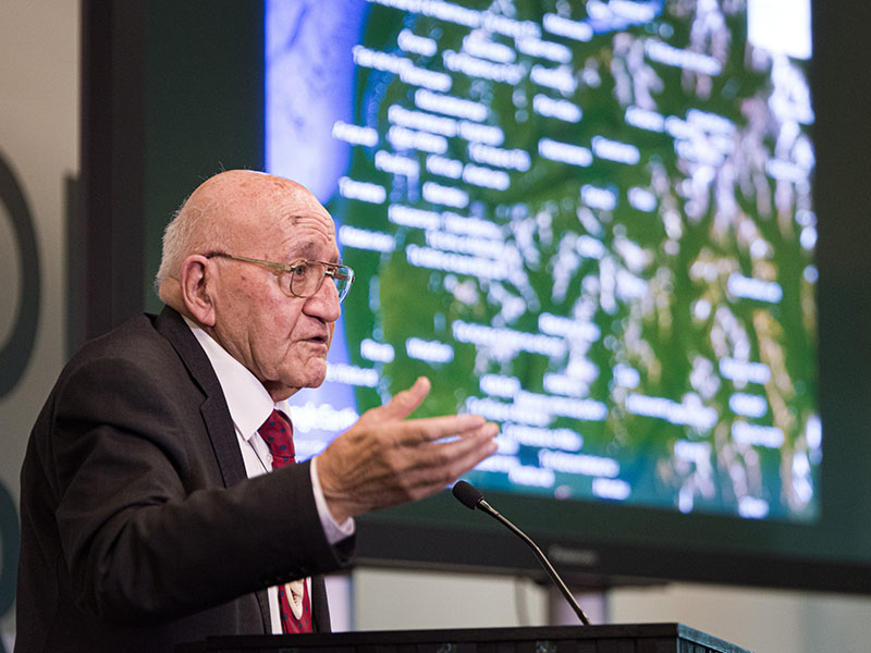 Ta Tipene addresses an audience in front of a large screen