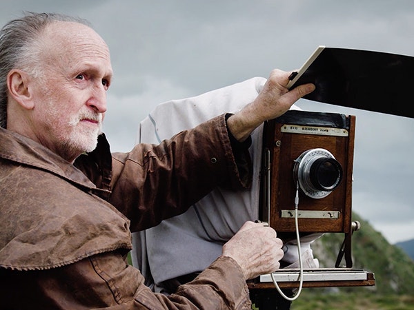 Mark Adams, 2019. Te Papa Mark Adams in a brown jacket looks out towards the sea, while preparing his large camera. In the distance you can see the shoreline