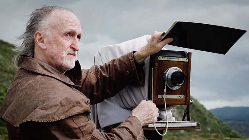 Mark Adams in a brown jacket looks out towards the sea, while preparing his large camera. In the distance you can see the shoreline