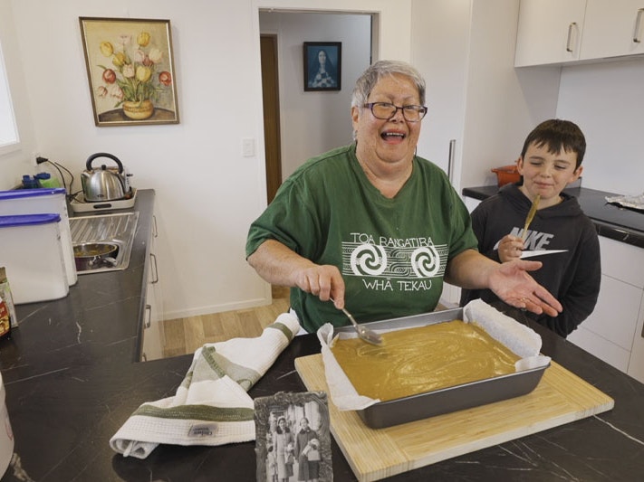 Still from Matariki kai: ginger crunch recipe, 2023. Te Papa Screenshot from a video, showing an older woman in a green T-shirt icing a cake in her kitchen as a young boy looks on