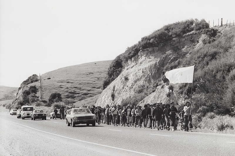 Black and white photo of a large group of people marching alongside a rural road. The person at the front is carrying a large flag