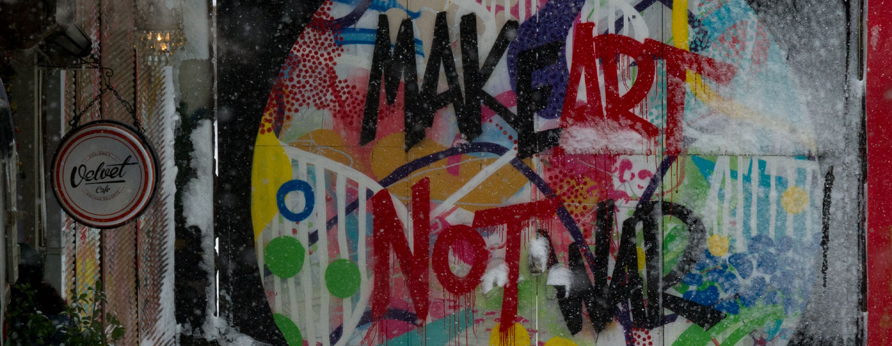 Photo of a wall with graffiti on it. The graffiti is a colourful circle and inside it is written "Make Art Not War". It has been snowing and snow has settled on the ground at the wall