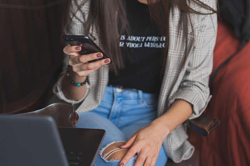 Close up photo of a woman sitting at a table, holding a phone, with a laptop open