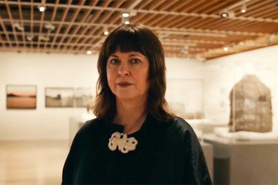 Head and shoulders of a woman with dark hair and dressed in black standing in a gallery