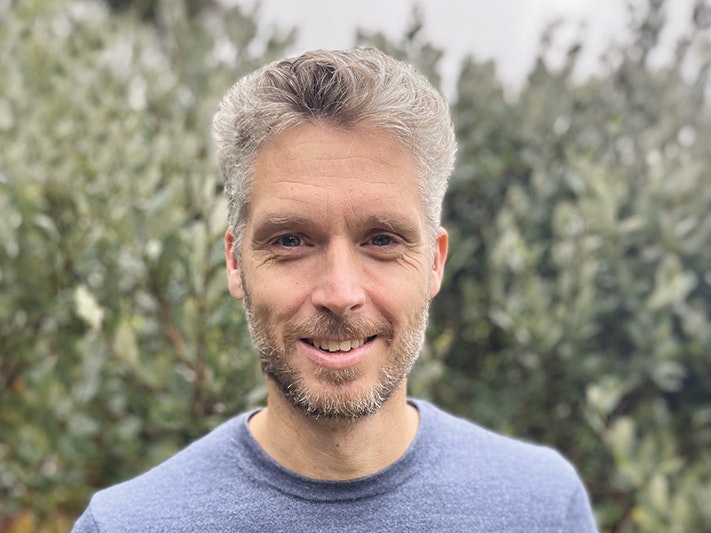 Ned Barraud. Photo courtesy of the author Head and shoulders of a white man with grey hair and a blue jumper. He is standing in front of green shrubbery.