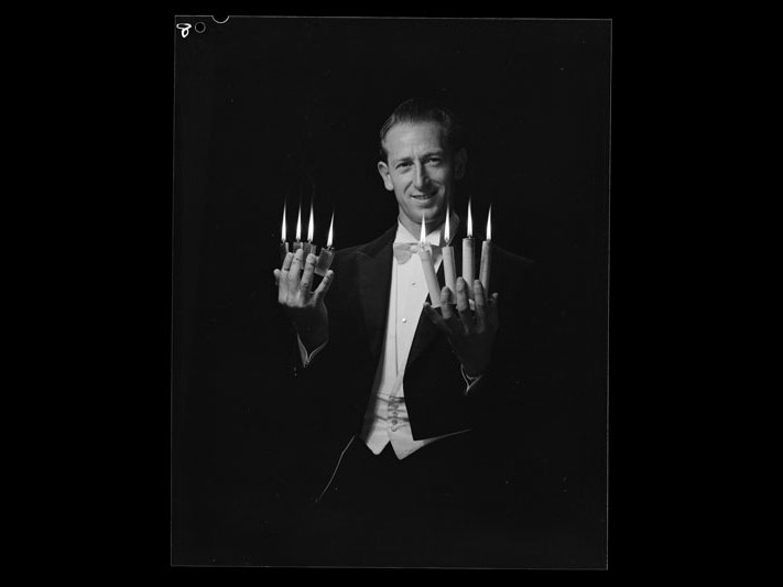 Magicians Graham Grant and Val, by Spencer Digby Studios, 29 March 1966, black and white film negative. Spencer Digby - Ronald D Woolf Collection. Gift of Ronald Woolf, 1975. Te Papa (B.081478) A magician poising for a photo. In his left hand area lit cigarette lighters between each finger. In his right handle, lit candles are held between each finger