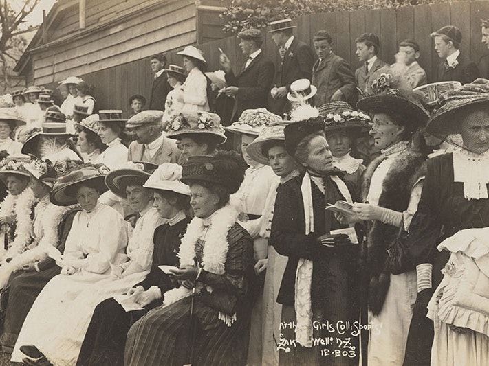 At the Girls College sports, about 1910, Wellington, by Zak (Joseph Zachariah). Te Papa (PS.003374) A large group of women sitting watching something out of frame. Men stand behind them