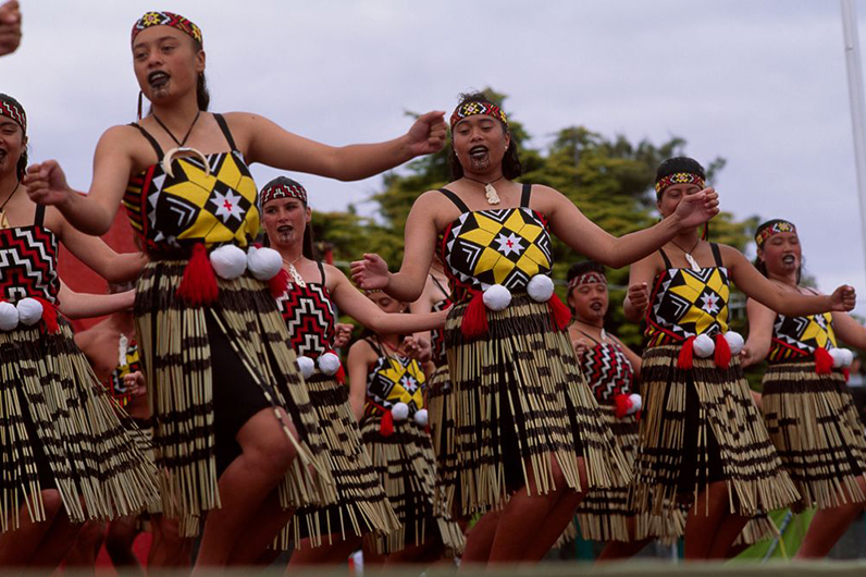 Women in traditional costume wearing white balls (poi) at their waists are singing and dancing in unison.