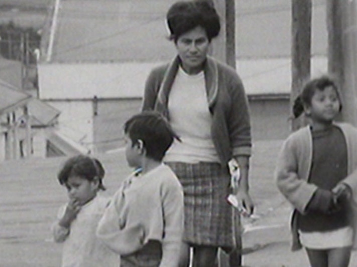 Still from Dawn Raids. NZ On Screen Black and white still from a video, showing a Polynesian women and three children walking down a street