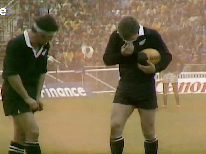 Still from ‘Re:visit - Remembering the Springbok Tour’ (Re: News/YouTube) Two members of the All Blacks huddle to avoid the smoke on the pitch from the flare