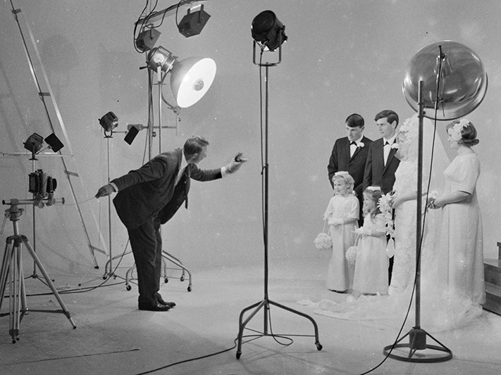 Crop of Christopher Bede Studio, by Frank Hofmann. Gift of the Frank Hofmann Estate, 2016. Te Papa (F.013805) A photographer has a puppet on one hand to entertain children while his other hand is operating a camera to take photos of a wedding party. The room is surrounded in lights and props.