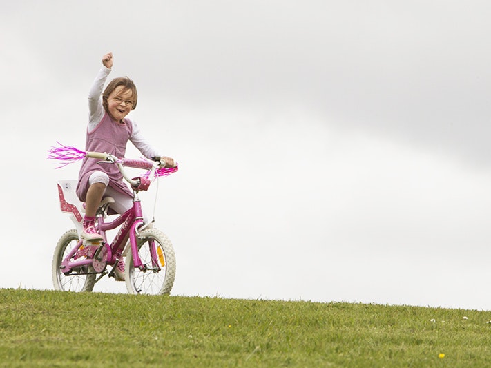 Maya Shaw, 6 years old, 2015. Photo by Norm Heke. Te Papa A child in a pink and white top riding a bright pink pushbike with pink tassells over grass and is punching the air with her right fist.